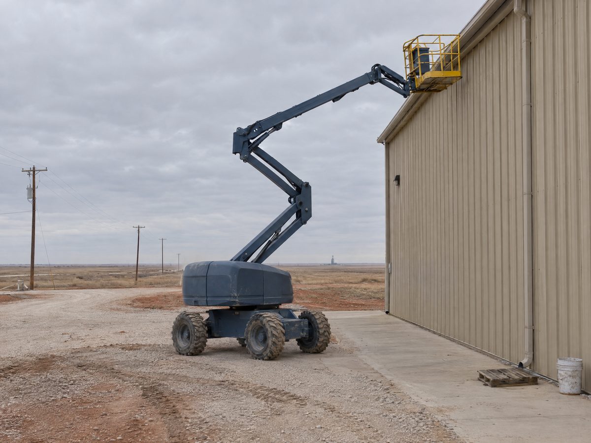 A blue-gray articulating boom lift raises its empty yellow-railed platform to the eave of a tall tan metal warehouse wall.