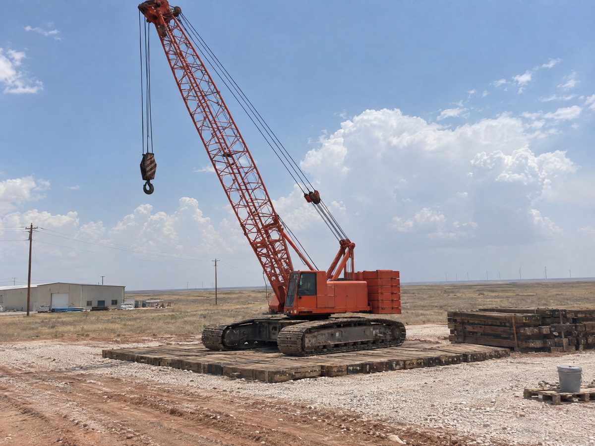 An orange-red lattice-boom crawler crane stands on heavy timber crane mats on a flat gravel site, with wind turbines far off on the horizon.