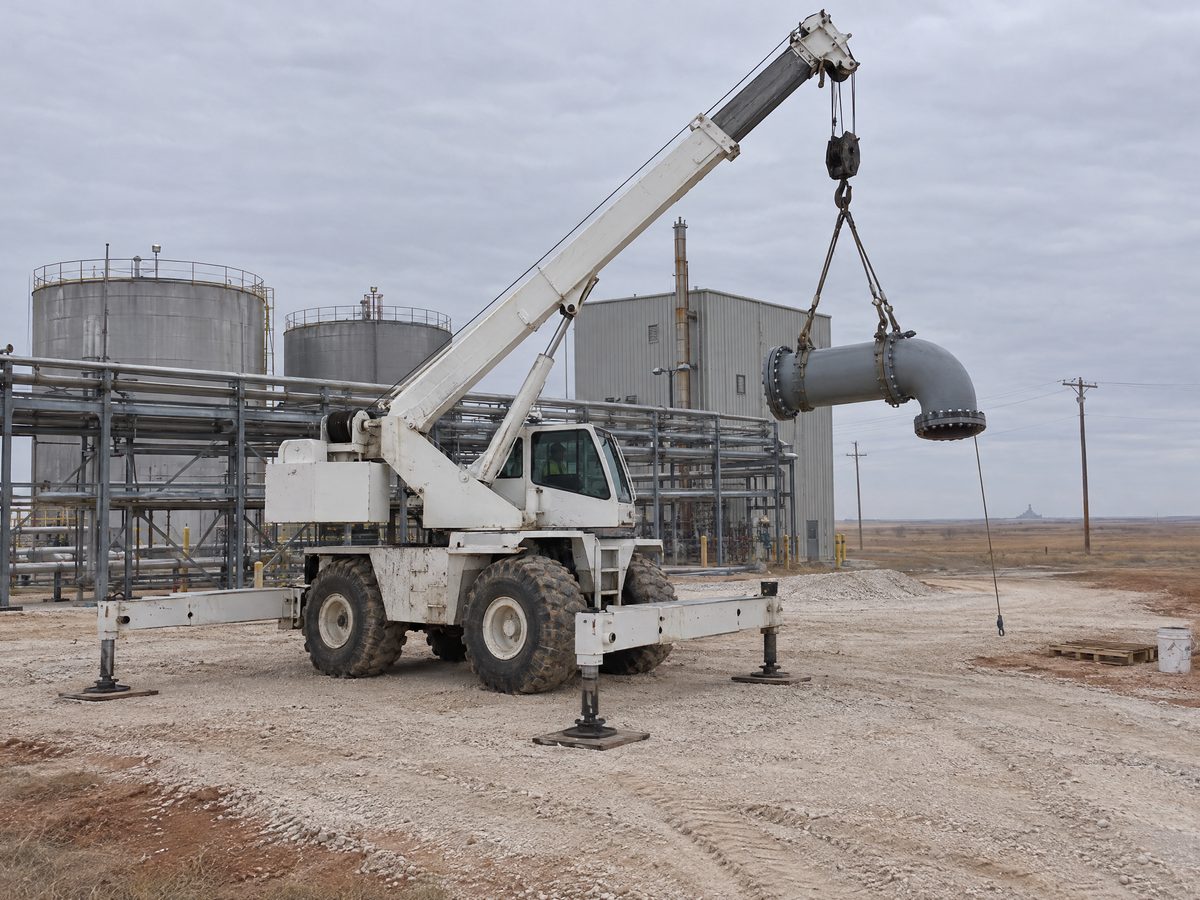 A white rough-terrain crane on outrigger pads lifts a flanged steel pipe spool with an elbow beside pipe racks and storage tanks at a plant.