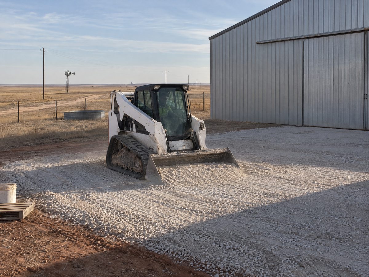 A white compact track loader spreads pale caliche gravel for a new pad in front of a metal barn on open ranch land.