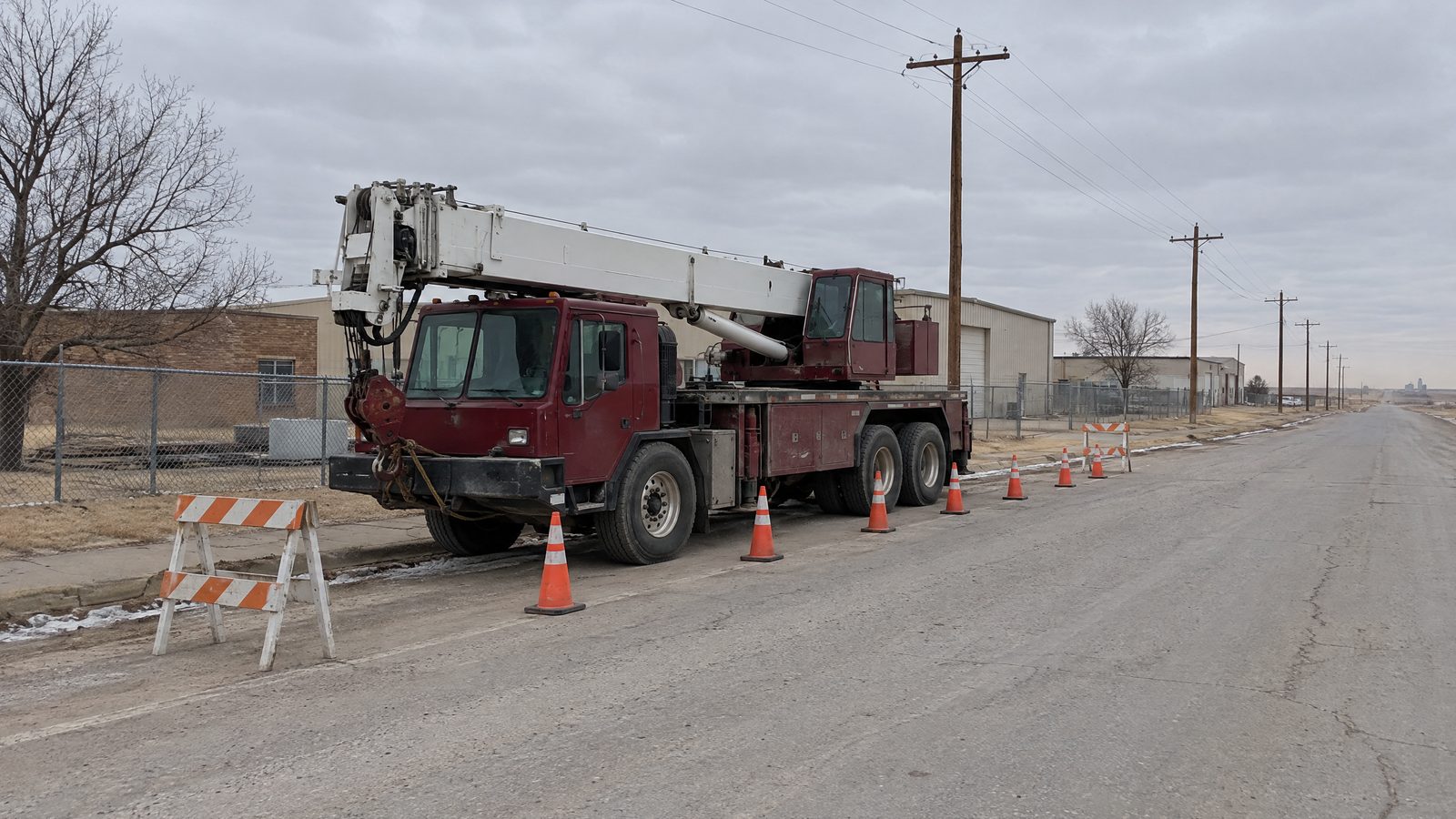 A dark red truck crane with its boom stowed sits at the curb of a wide industrial street, ringed by cones and striped barricades.
